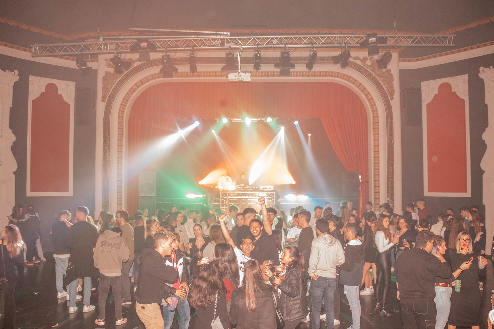 Fiesta con gente bailando con escenario al fondo y un DJ junto a mesa de mezclas