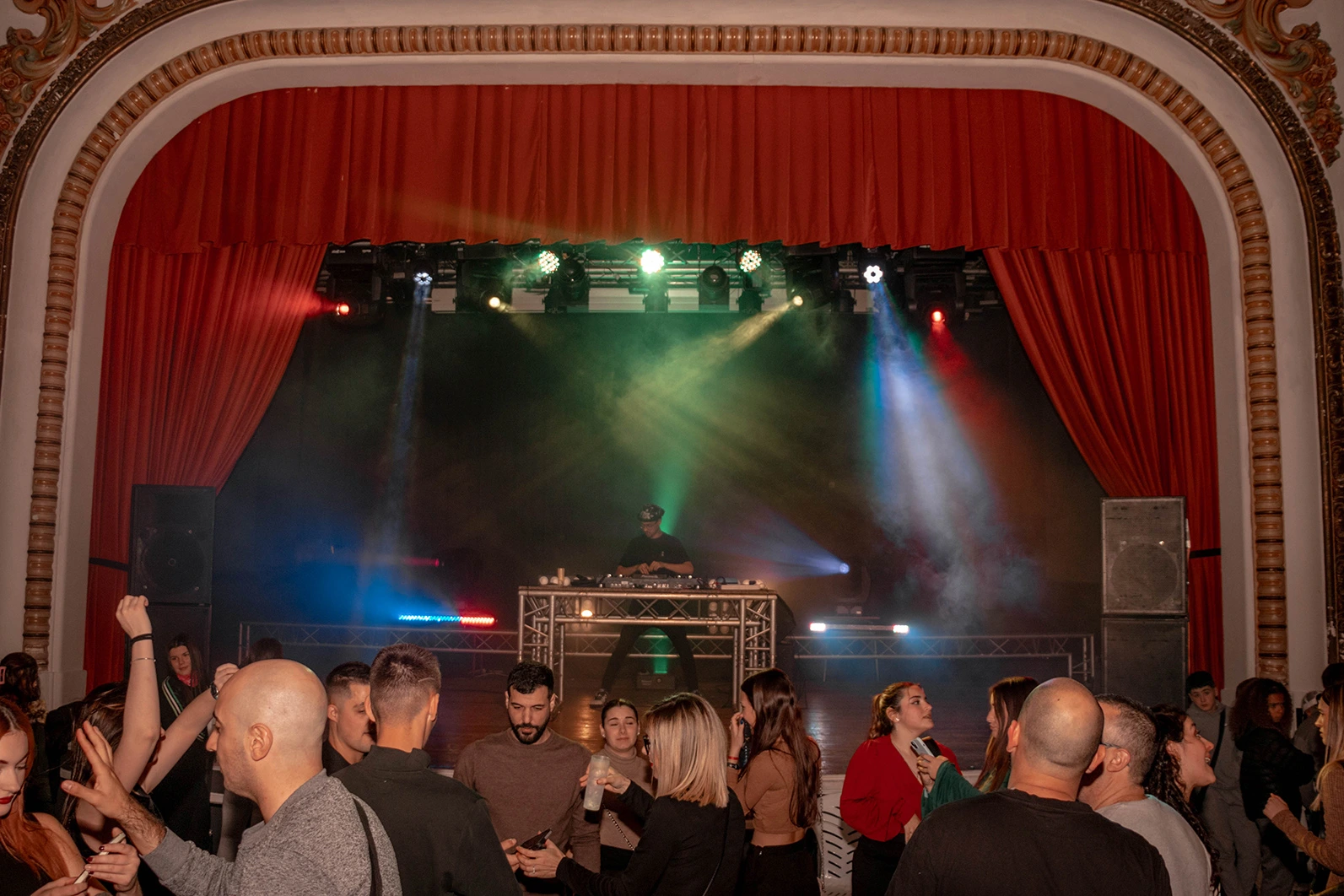 Fiesta con gente bailando con escenario al fondo y un DJ junto a mesa de mezclas